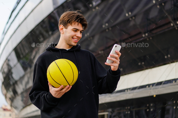 Young basketball fan in hoodie holding ball and watching match on smartphone. Happy man win a bet.