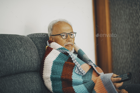 One old senior man sitting on the sofa at home with colorful wool cover for low temperature cold flu