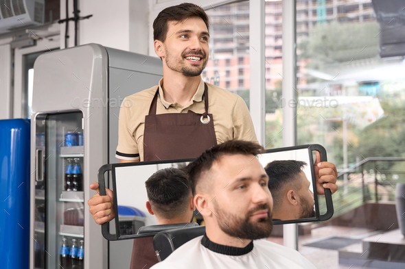Contented hairstylist helping guest to see his haircut in mirror