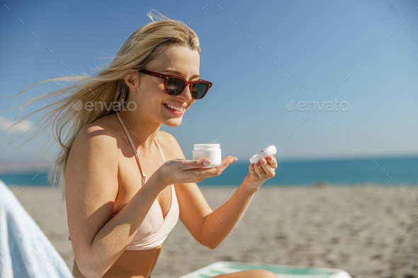 Excited lady in sunglasses opening a jar with body cream while relaxing on beach