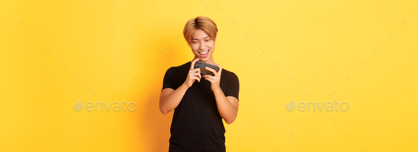 Portrait of stylish handsome asian guy playing smartphone game, looking amused at mobile screen