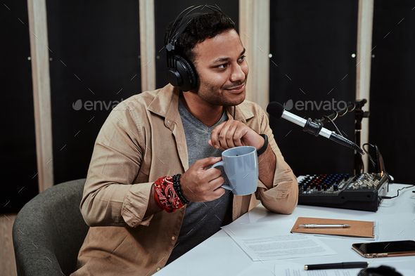 Portrait of young male radio host going live on air, talking with guest, holding a