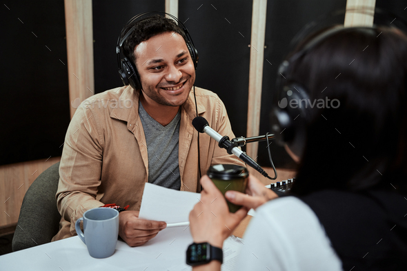 Portrait of young male radio host going live on air, talking with female guest, holding