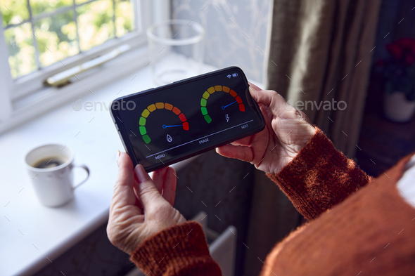 Senior Woman With Smart Meter Trying To Keep Warm By Radiator During Cost Of Living Energy Crisis