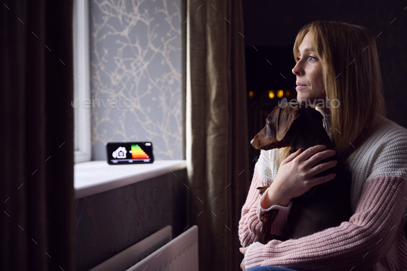 Woman With Dog And Smart Meter Trying To Keep Warm By Radiator In Cost Of Living Energy Crisis