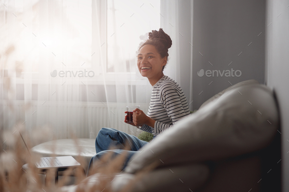 Pretty mixed-race young woman in good mood chilling on couch at cozy home with