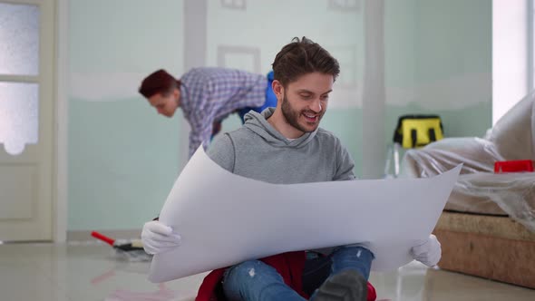 Thoughtful Concentrated Man Examining Blueprint Sitting on Floor with Blurred Woman Painting Wall at alt