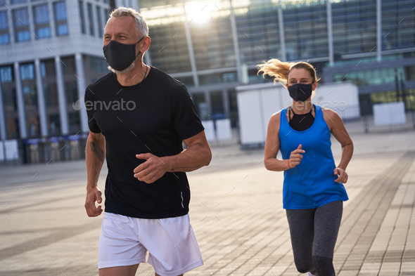 Sporty middle aged couple wearing protective masks running in urban environment, staying active