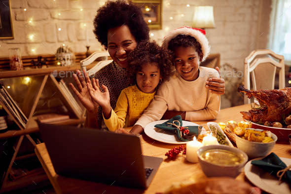Happy black mother and kids waving during video call on Christmas day at dining table.