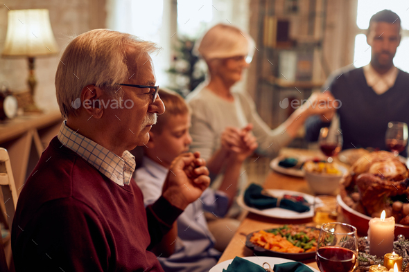 Senior man and his family saying grace while having Thanksgiving dinner at dining table.