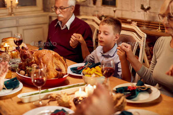 Small boy and his family holding hands and praying before Thanksgiving meal at dining table.