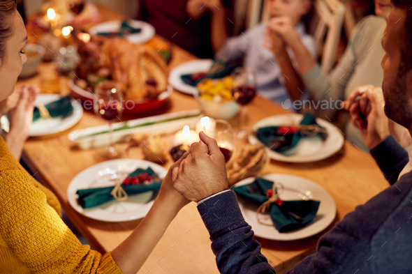 Close-up of extended family holding hands and praying before Thanksgiving meal at dining table,
