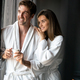 Happy young couple in white bathrobes drinking coffee together. Hotel, travel, relationships concept - PhotoDune Item for Sale