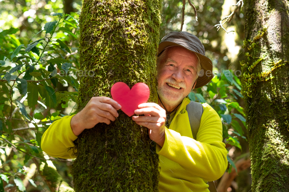 Smiling senior man partially hidden by a moss covered tree trunk in the woods holding a paper heart