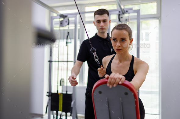 Physical instructor assisting young woman working out on pull cable exercising machine