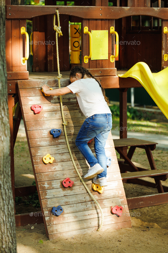 Little girl climbing rope and step up sports game complex in playground.  Summer holidays in camp Stock Photo by burmistrovaiuliia