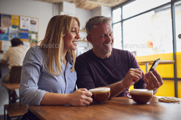 Couple Sitting At Table In Coffee Shop Looking At Mobile Phone Together - Stock Photo - Images