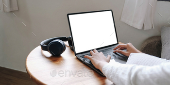 Girl typing on laptop with blank screen, headphones on wooden table, mock up
