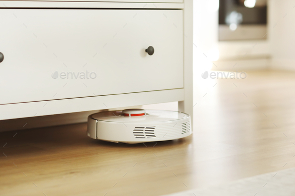 White robotic vacuum cleaner on laminate floor cleaning dust in living room interior. Smart