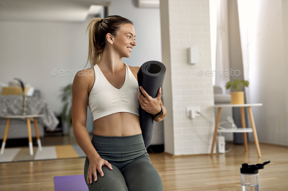 Young happy female athlete with exercise mat in the living room.