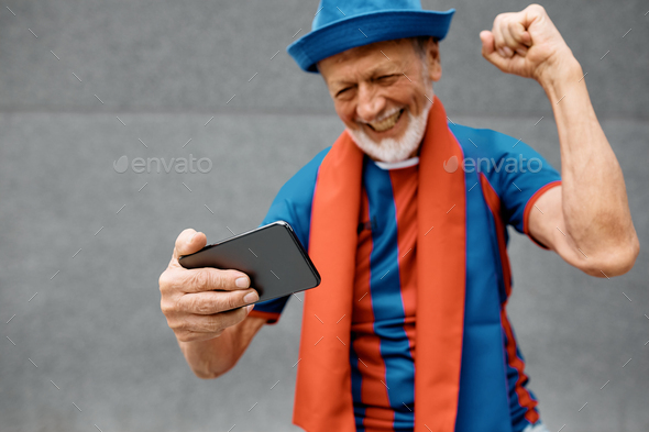 Close up of senior soccer fan cheering while watching a game on mobile phone.