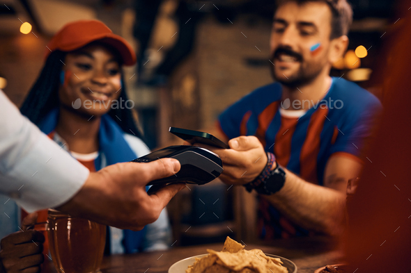 Close up of couple of sports fans using smart phone and paying contactless in bar.