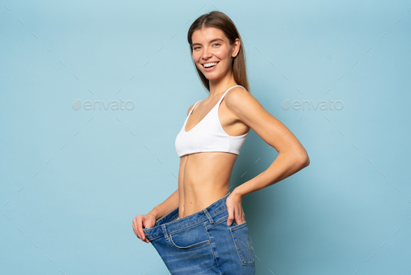 Side view of slim woman in big jeans showing her diet results and weight loss on blue background