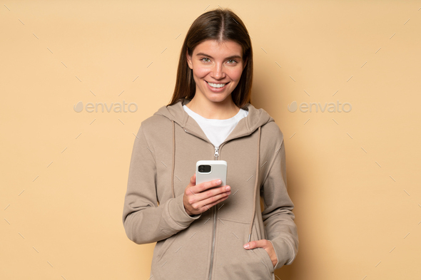 Woman in hoodie downloading fitness app to her phone to train in morning isolated on brown - Stock Photo - Images
