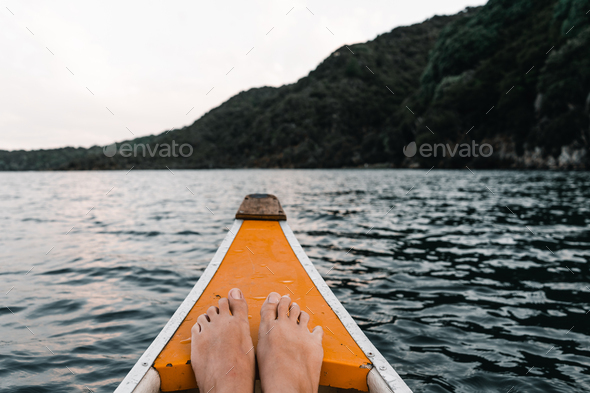 feet together resting on the prow of a white and orange wooden canoe heading into the forest
