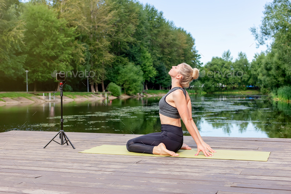 A woman in a on a wooden platform in summer, does yoga on green mat by pond