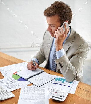 Shot of a young businessman talking on the phone while writing at his desk