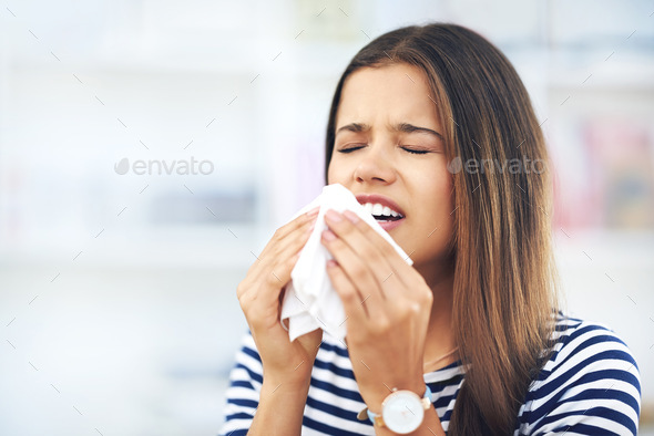 Achooooo. Shot of a young woman with allergies sneezing into a tissue at home.