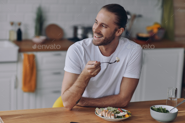 Cheerful young man enjoying lunch while sitting at the kitchen counter at home