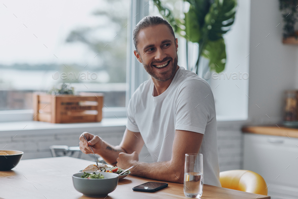 Joyful young man having lunch while sitting at the kitchen counter at home