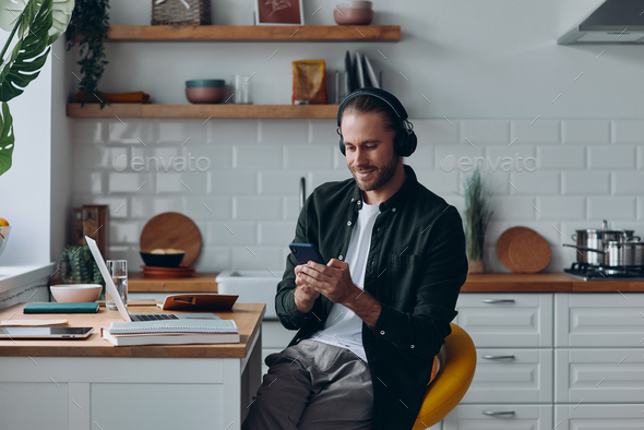 Confident man in headphones using smart phone while sitting at the kitchen counter at home