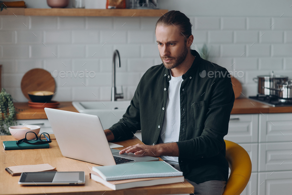Confident young man using laptop while working at the kitchen counter at home