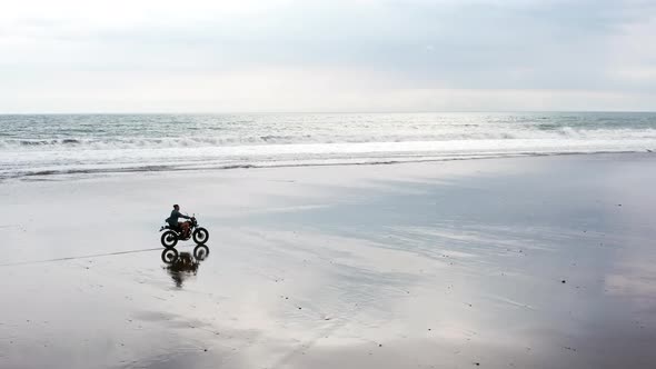 Young Handsome Hipster Man Riding Modern Custom Motorcycle Racer on the Black Sand Beach alt