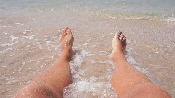 Closeup of a Man's Legs Lying on a Sandy Beach in Azure Sea Water Small Waves Beating Against His