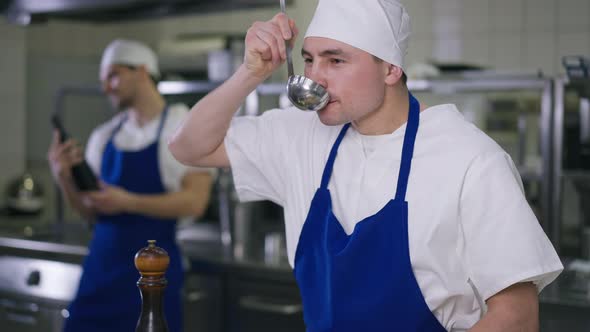 Caucasian Chef Tasting Soup Adding Salt to Dish Standing in Kitchen with Cook Preparing Food at alt