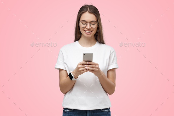 Young woman looking at phone, smiling while holding it in both hands, isolated on pink background - Stock Photo - Images