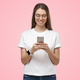 Young woman looking at phone, smiling while holding it in both hands, isolated on pink background - PhotoDune Item for Sale