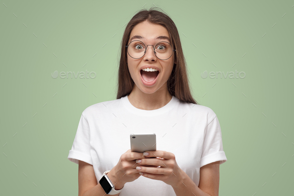 Close-up of girl in white t-shirt, holding phone, looking surprised and shocked, isolated on green - Stock Photo - Images