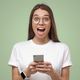 Close-up of girl in white t-shirt, holding phone, looking surprised and shocked, isolated on green - PhotoDune Item for Sale