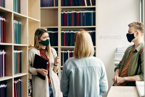 Smiling university student and her friends wearing face masks while communicating in a library.