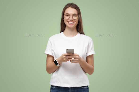 Portrait of attractive young female in white t-shirt, holding smartphone, smiling at camera - Stock Photo - Images