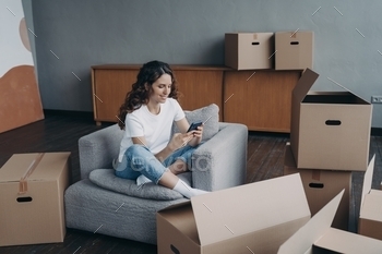 Girl uses phone app, orders moving company, shopping online for new home, sitting with carton boxes
