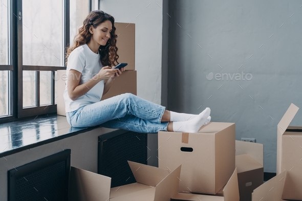 Happy girl using smartphone apps, choosing moving company, sitting with boxes. Relocation, new home