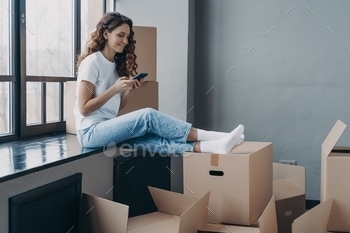 Happy girl using smartphone apps, choosing moving company, sitting with boxes. Relocation, new home