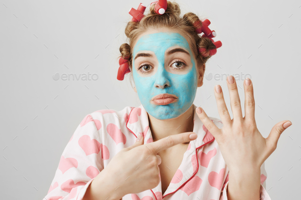 Portrait of upset funny housewife in hair-curlers pyjamas and blue face mask pointing at left arm