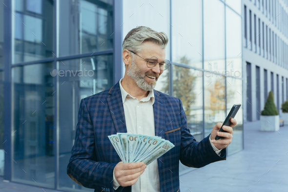 Happy mature gray-haired businessman holding winnings money cash in hands, scared man smiling and